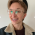 A person with short hair and glasses wearing a dark top and jean jacket is smiling